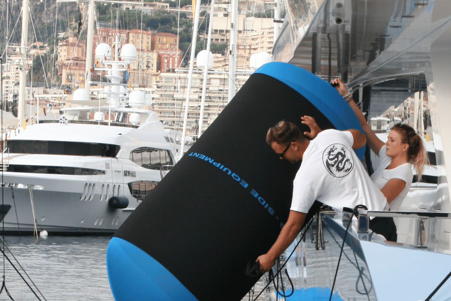 Two people interacting with a large blue boat fender and black fender cover on a yacht with yachts in the background.